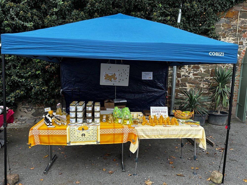 Marktstand Bienen-Oase mit blauem Zelt, orange gedecktem Tisch, Honiggläsern und Waben-Herz-Schmuck